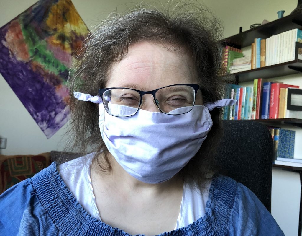 Kopf und Schultern von Flora. Sie trägt eine Brille und einen Mund-Nasen-Schutz
