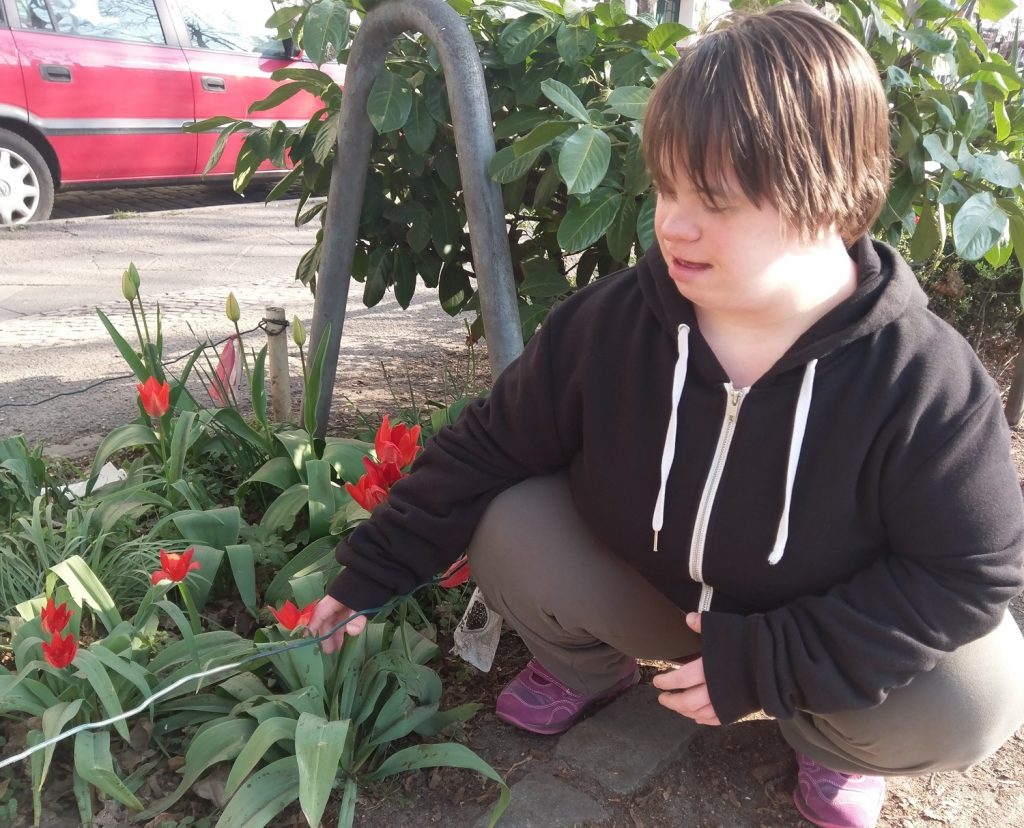 Marion hockt vor einem Blumen-Beet mit roten Tulpen und berührt eine Tulpe. Im Hintergrund die Straße. Ein rotes Auto parkt gegenüber.