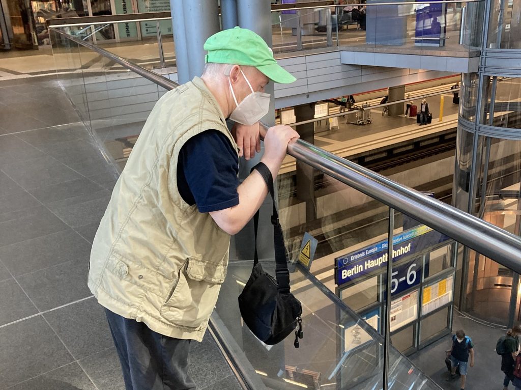 Frank lehnt an ein Geländer und schaut hinunter auf den Bahnsteig. Er trägt eine grüne Mütze und einen Mundschutz. Auf dem Schild auf dem Beinsteig steht Hauptnbahnhof.