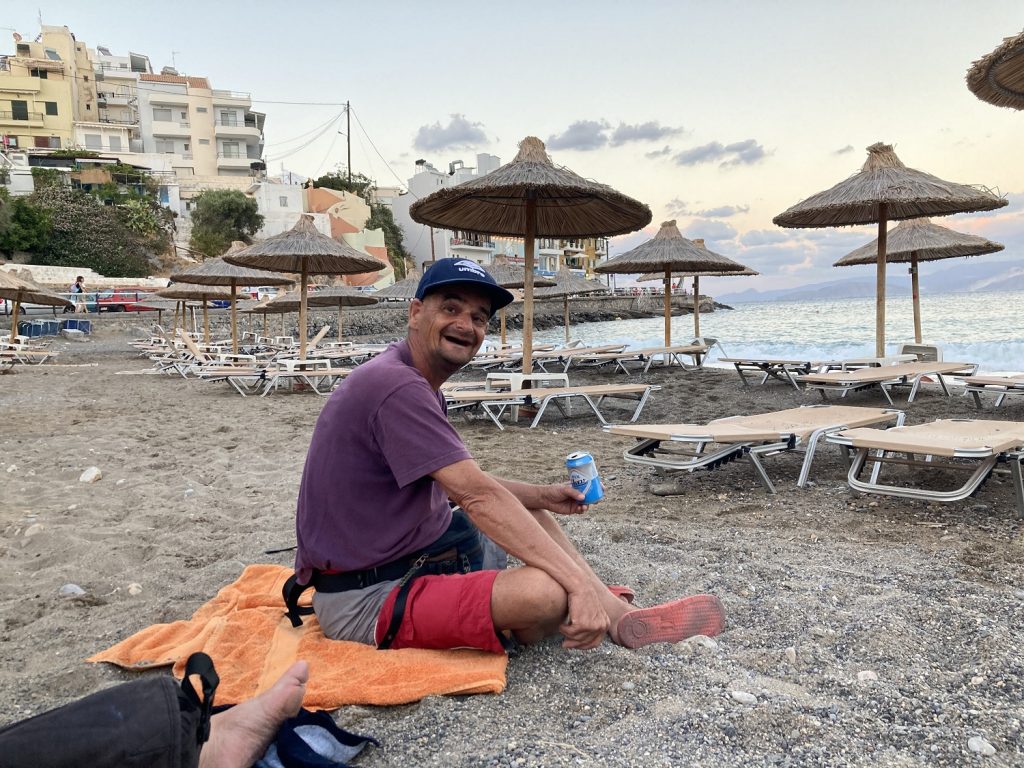 Matti sitzt am Strand. Im Hintergrund sind Sonnenschirme und Liegen zu sehen und das Meer.d