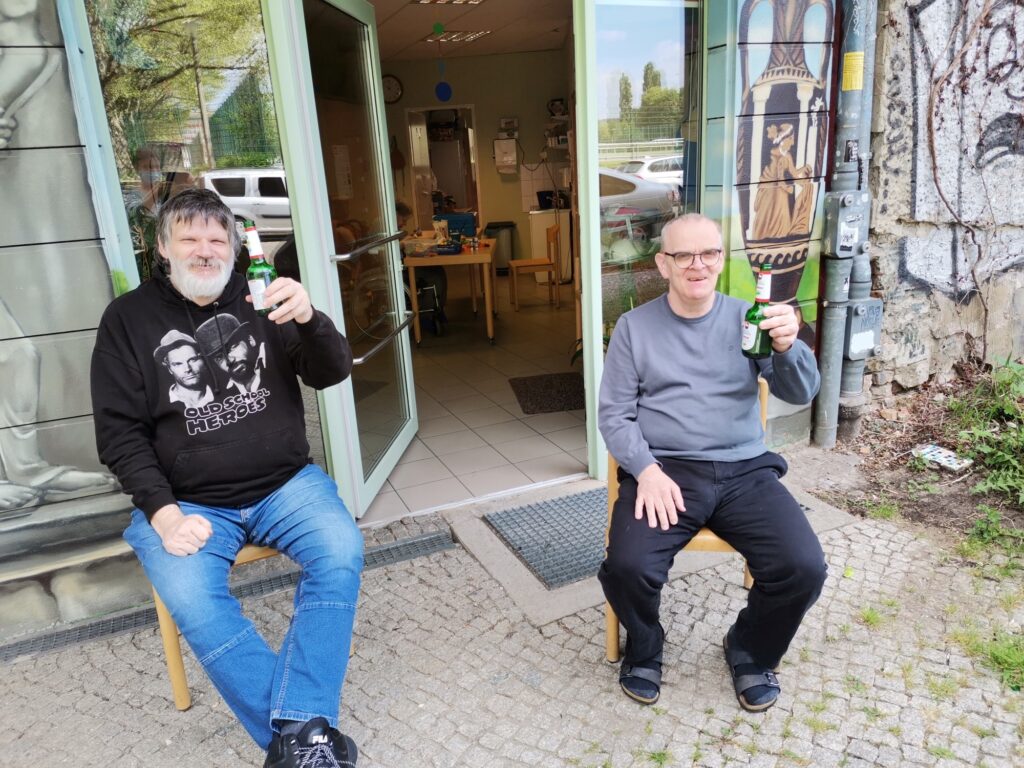 René und Michael sitzen auf Stühlen auf dem Gehweg vor der WoZerei