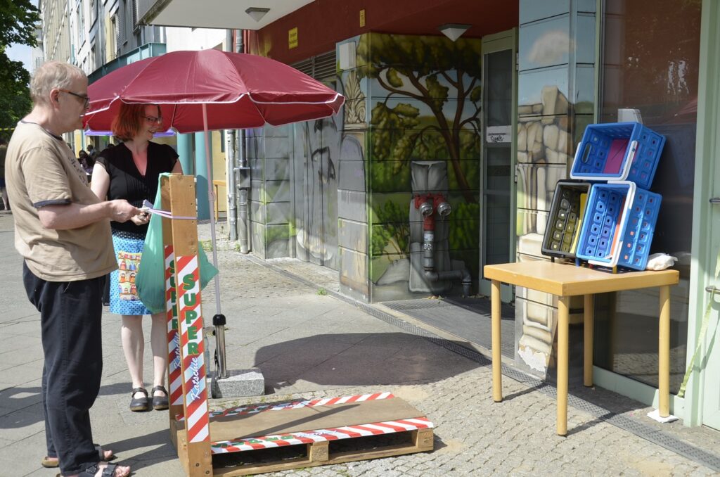 Michael und eine Frau stehen bei einem Sonnenschirm. Michael schießt Papierschnipsel in Richtung eines Tisches mit drei Plastikboxen..