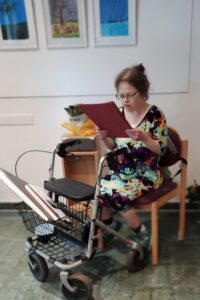 Flora sitzt auf einem Stuhl, ihren Rollator vor sich. Sie liest ihre Rede vor. Im Hintergrund hängen ihre Biulder an der Wand.
