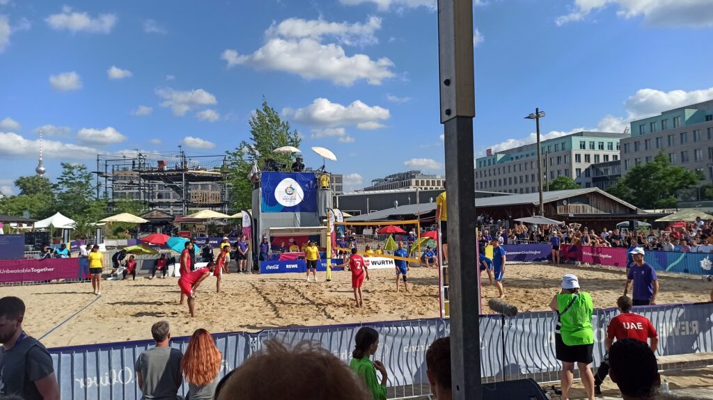 Ein BEachvolleyballfeld. Es findet gerade ein Spiel statt. Das Wetter ist schön, im Hintergrund blauer Himmel.