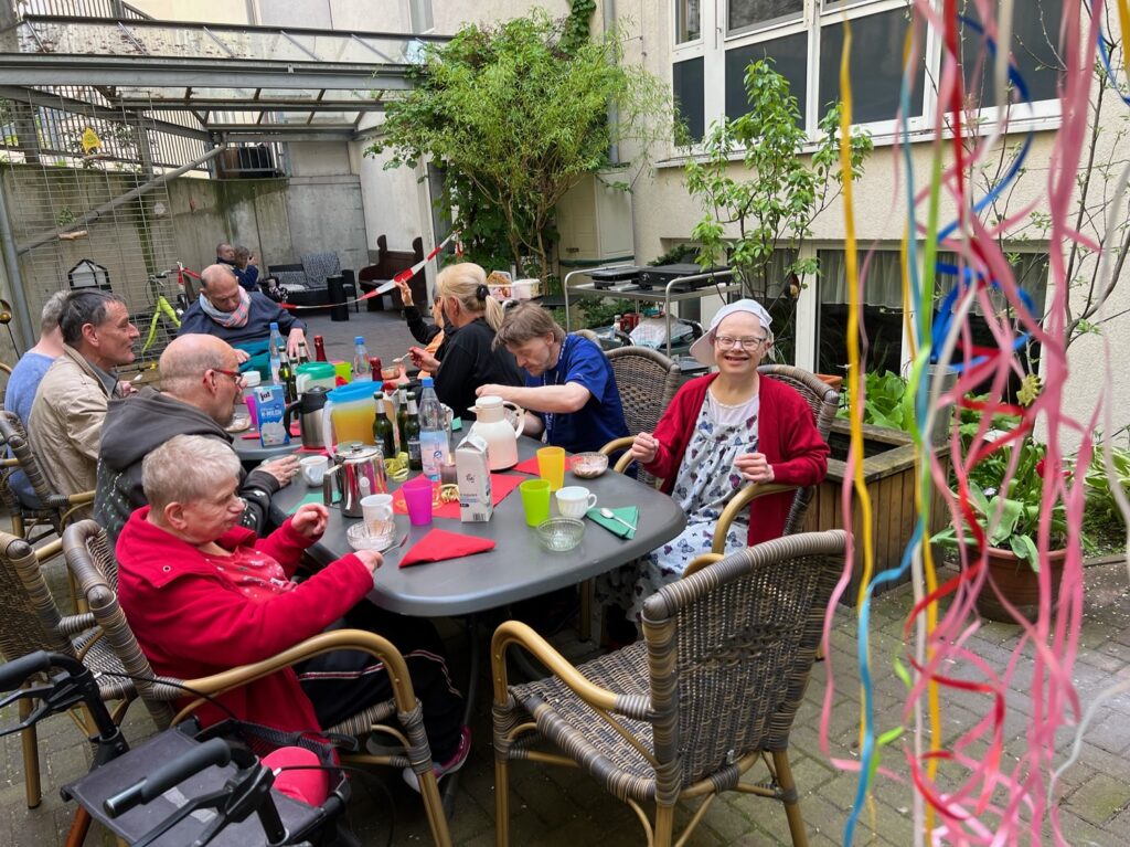 Eine Gruppe Menschen sitzt um einen Tisch in einem Hof. Der Tisch ist gedeckt. Rechts hängen Luftschlangen herunter.