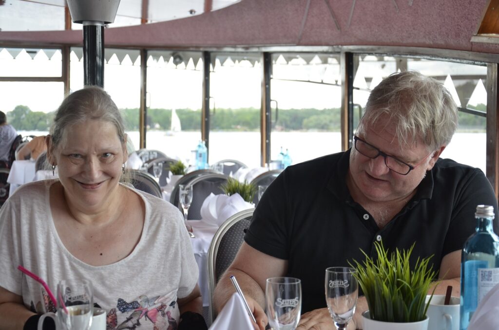Eine Frau und ein Mann sitzen nebeneinander an einem Tisch. Der Mann schreibt. Die Frau lächelt.