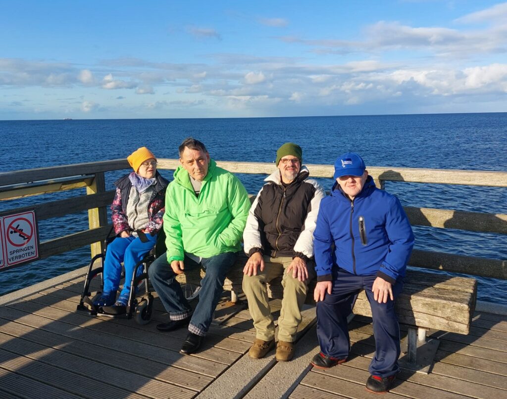 Eine Frau und drei Männer sitzen nebeneinander auf einer Seebrücke, die Frau im Rollstuhl, die drei Männer auf einer Bank. Im Hintergrund ist die Ostsee zu sehen. Der Himmel ist blau bis auf ein paar Wolken. Die vier tragen Jacken.