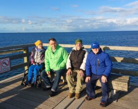 Eine Frau und drei Männer sitzen nebeneinander auf einer Seebrücke, die Frau im Rollstuhl, die drei Männer auf einer Bank. Im Hintergrund ist die Ostsee zu sehen. Der Himmel ist blau bis auf ein paar Wolken. Die vier tragen Jacken.
