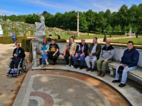 Das Bidl zeigt eine Gruppe Menschen, die auf einer langen gebogenen und setinernen Bank vor den Terrassen von Schloss Sanssouci in Potsdam sitzen. Das Schloss selbst ist nicht zu sehen. Die Menschen sitzen nebeneinander, einer sitzt in einem Rollstuhl neben der Bank.