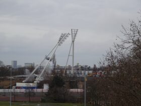Zwei große weiße Flutlichtmasten. Einer der Masten ist dabei umzukippen, Rauch steigt auf. Bagger und Schutt sind unten zu sehen. Im Hintergrund ist eine Stadtkulisse zu sehen, der Himmel ist bewölkt.