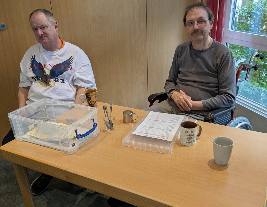 Klaus und Norbert sotzen an einem Tisch, Norbert hat ein Blat vor sich liegen, Klaus eine durchsichtige Plastikbox. Es stehen Tassen auf dem Tisch.
