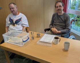 Klaus und Norbert sotzen an einem Tisch, Norbert hat ein Blat vor sich liegen, Klaus eine durchsichtige Plastikbox. Es stehen Tassen auf dem Tisch.
