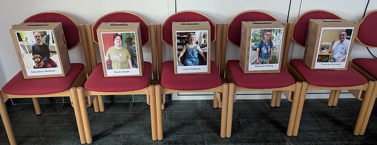 Fün Stühle mit roten Polstern stehen aufgereiht an einer Wand. Auf den Stühlen stehen Boxen aus Karton. Die Boxen sind mit jeweils einem Foto beklebt. Unter den Fotos stehen Namen.