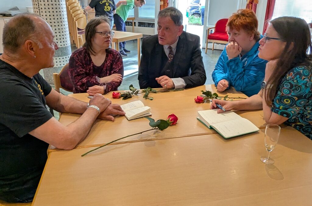 Eine Gruppe von fünf Personen sitzt an einem Holztisch und bespricht sich. Es liegen rote Rosen und Notizbücher auf dem Tisch.