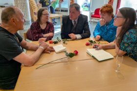 Eine Gruppe von fünf Personen sitzt an einem Holztisch und bespricht sich. Es liegen rote Rosen und Notizbücher auf dem Tisch.