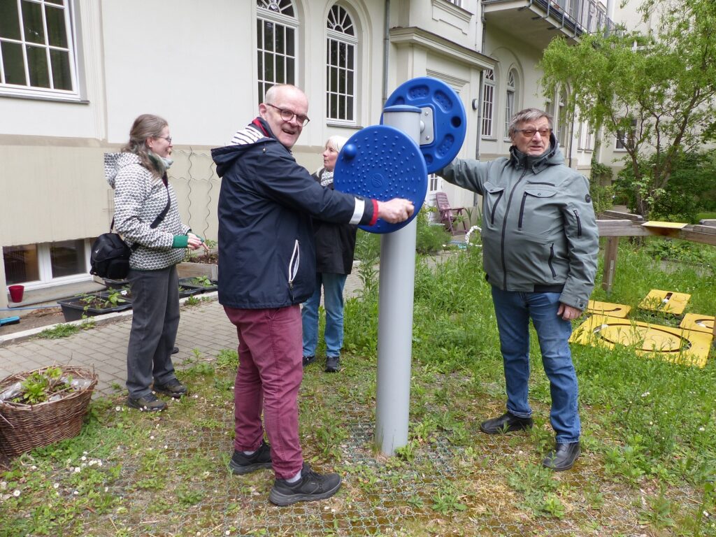 Zwei Männer stehen in einem Außenbereich vor einem Gebäude und benutzen ein Trainingsgerät. Zwei Frauen sind im Hintergrund zu sehen. Es ist ein bewölkter Tag. Alle vier tragen Jacken. Das Trainingsgerät in der Mitte ist ein vertikaler Pfosten mit zwei blauen, runden Scheiben, die mit kleinen Noppen übersät sind. An den Scheiben sind Griffe zum Drehen. Die beiden Männer drehen die Scheiben.